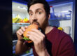 Man eating burger near refrigerator in kitchen at night Bad habit