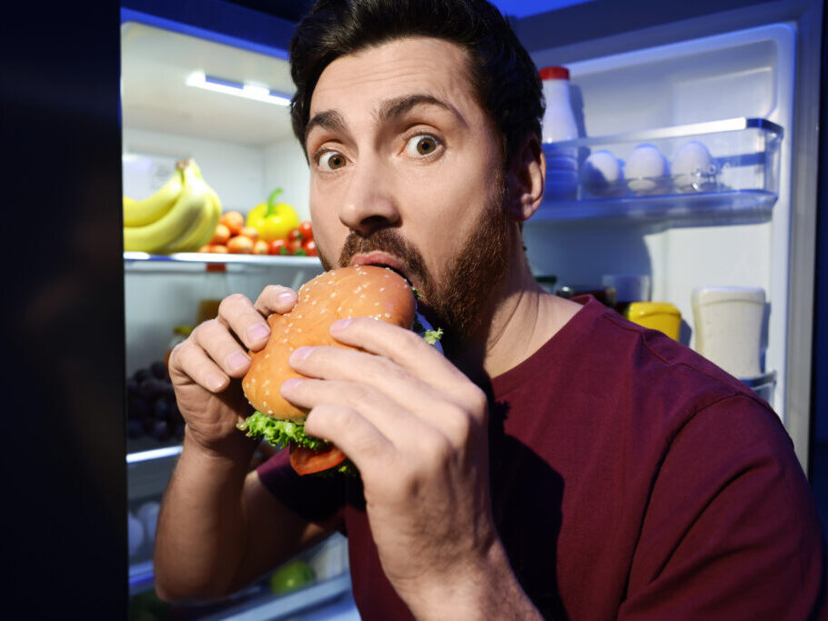 Man eating burger near refrigerator in kitchen at night Bad habit Man eating burger near refrigerator in kitchen at night Bad habit